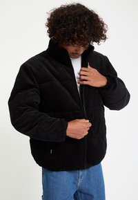 Homme aux cheveux bouclés fermant une doudoune noire en velours côtelé, portant une chemise blanche et un jean bleu sur un fond uni.
