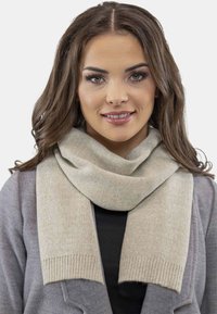Écharpe beige clair en tissu doux, avec une texture lisse et des extrémités côtelées, drapée autour du cou sur un haut sombre et un manteau gris.
