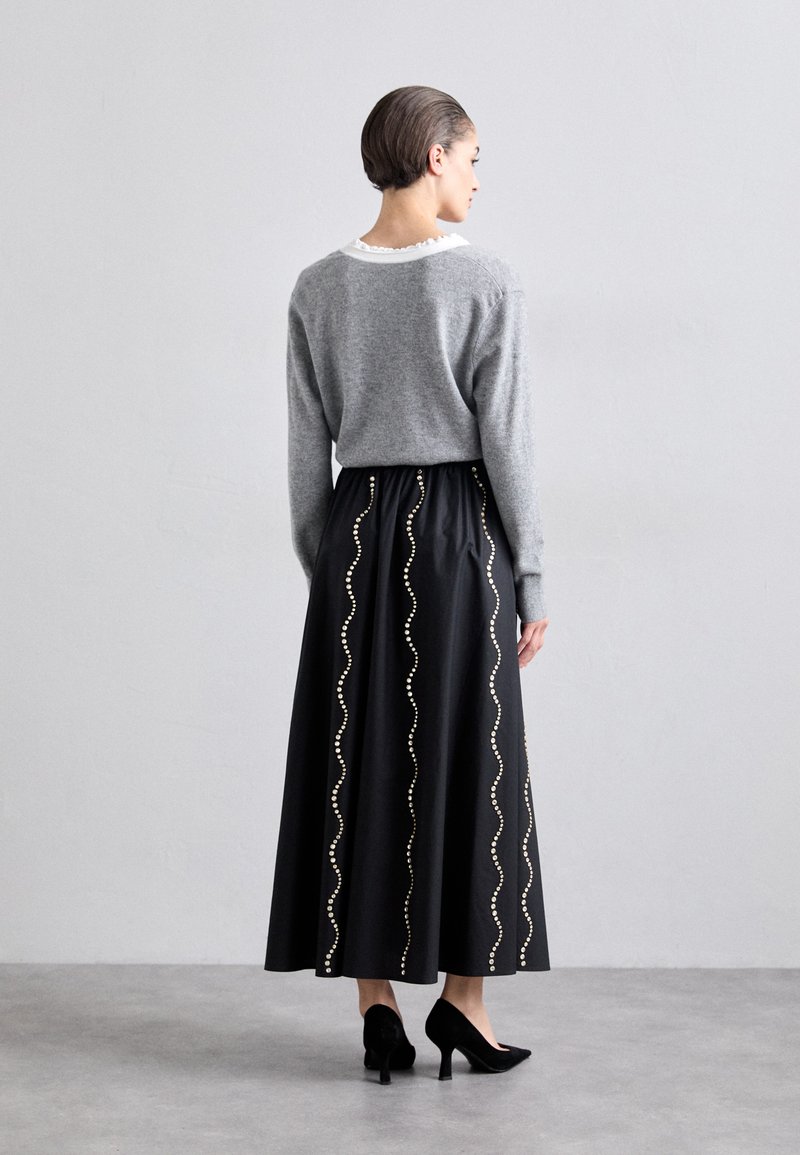 Grauer Pullover mit weißem Kragen, kombiniert mit einem schwarzen Maxirock mit wellenförmigen goldenen Akzenten. Modell trägt schwarze Absätze, von hinten gesehen.