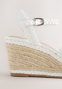 Sandale compensée en cuir tressé blanc avec bride de cheville et talon plateforme en corde tressée naturelle, détaillée avec une boucle argentée.