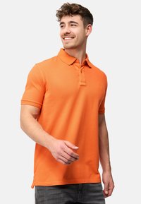 Narančasta polo majica s ovratnikom, kratkim rukavima i suptilnom teksturom. Ima placket s tri gumba i mali logo na prsima.