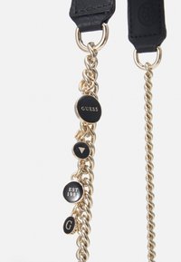 Svart läderrem med guldkedjeaccentuationer, med runda taggar med "GUESS," "EST 1981," och andra logotyper. Slät textur, modern design.