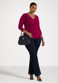 Fuchsia omslagsbluse med trekvart ermer, kombinert med mørke vide jeans. Modellen holder en marineblå teksturert håndveske med klaff og sølvdetaljer.