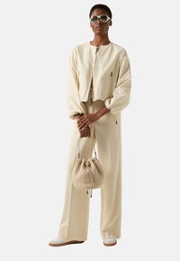 Krótkja kremowa kurtka z bufiastymi rękawami, pasujące szerokie spodnie oraz beżowa torba ze sznurkiem. Modelka ma na sobie okrągłe okulary przeciwsłoneczne.