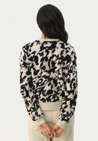 Femme aux longs cheveux noirs portant un pull à motif abstrait noir et blanc, debout avec les mains jointes dans le dos.