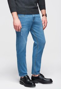 Jean bleu clair avec un léger effet usé, associé à des chaussures en cuir noir. La tenue est complétée par un pull côtelé gris foncé.