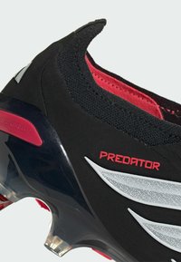 Κοντινό πλάνο μαύρου και κόκκινου ποδοσφαιρικού παπουτσιού Predator με μαύρο πλεκτό κολάρο, κόκκινη επένδυση, ασημένιες λεπτομέρειες και διάφανα καρφιά σε μαύρη σόλα.