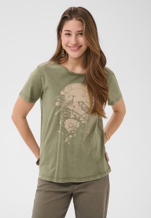 Ung kvinde med langt brunt hår, smilende, iført en olivengrøn t-shirt med et design af en fugl og en rose, kombineret med brune bukser.