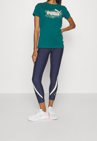 T-shirt in cotone teal con grafica floreale bianca; leggings blu navy con dettagli bianchi e verdi; sneakers bianche con suola rosa.
