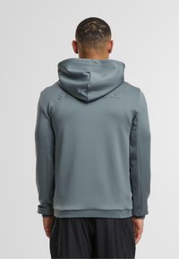 Šedá mikina s kapucí a hladkým povrchem, s logem v zdviženém písmu na zadní části a síťovinovými panely po stranách.