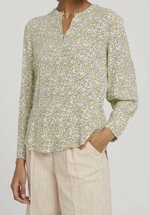 Femme portant un chemisier blanc à manches longues avec un petit motif floral jaune et un pantalon beige à jambes larges, posant avec une main dans la poche.