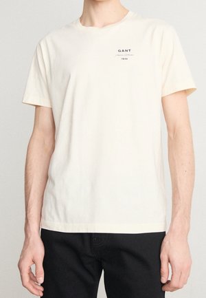 Osoba ubrana w gładki kremowy T-shirt z krótkim rękawem z małym napisem "GANT 1949" na lewej górnej części klatki piersiowej, w połączeniu z czarnymi spodniami.