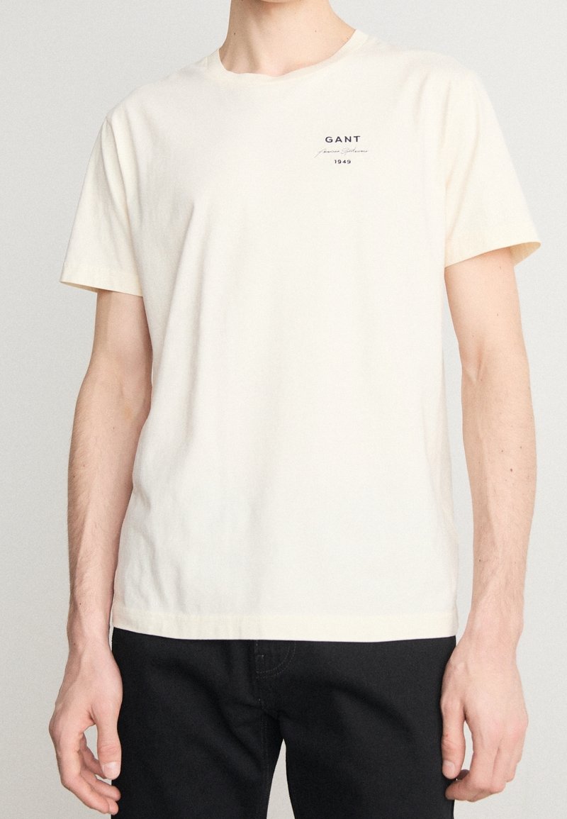 Personne portant un T-shirt uni crème à manches courtes avec un petit texte « GANT 1949 » sur le haut à gauche de la poitrine, associé à un pantalon noir.