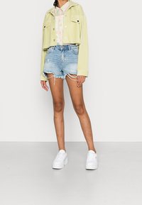 Veste en velours côtelé vert clair coupée avec deux poches avant, associée à une chemise à fleurs pastel, un short en denim déchiré et des baskets blanches à plateforme.