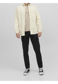 Ljusbeige skjorta med knappar över en randig t-shirt i brunt och krämfärg, kombinerat med svarta cargo-byxor och svarta sneakers med vita sulor.