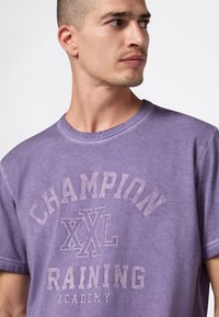 Mężczyzna w fioletowej koszulce z napisem "CHAMPION XXL RAINING ACADEMY", patrzy w prawo na tle białego, gładkiego tła.