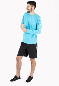 Lyseblå langærmet træningsskjorte med lynlås ved nakken. Sorte shorts med en sidelomme. Sorte træningssko med hvide såler.