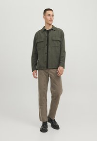 Giacca di pile verde con chiusura a bottoni, due tasche sul petto e dettagli neri, abbinata a pantaloni beige e stivali neri.