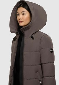 Padded jacket in dark gray with a large, structured hood, front zip closure, and logo on the sleeve. Features a smooth, textured finish.