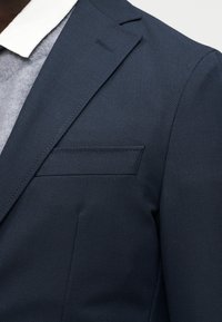 Κοντινό πλάνο ενός ατόμου που φοράει ένα σκούρο ναυτικό μπλε υφασμάτινο σακάκι πάνω από ένα γκρι πουλόβερ και λευκό πουκάμισο με γιακά.