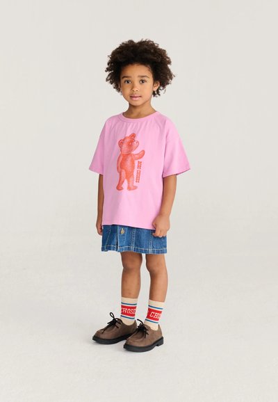 Enfant aux cheveux bouclés portant un t-shirt rose avec un motif d'ours rouge, une jupe en jean, des chaussettes rayées et des chaussures marron, debout sur un fond clair.
