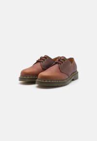 Braune Lederschuhe mit abgerundetem Zehenbereich, versehen mit flachen Schnürsenkeln, einem hellbraunen Innenfutter und einer strukturierten Gummisohle mit gelben Nähakzenten.