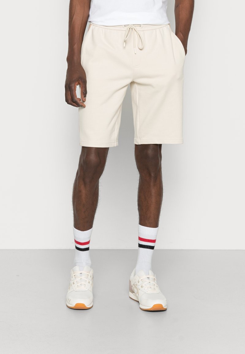 Homme portant un short beige à cordon, des chaussures de sport blanches et des chaussettes blanches montantes avec des rayures rouges et noires, debout devant un fond uni.