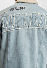 Chaqueta de mezclilla azul clara con cuello de borreguito blanco, letras grandes desgastadas "CARLK" y "EST. 1989" bordado en la parte posterior.