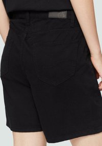 Shorts en denim noir, taille mi-haute, avec un design classique à cinq poches. Le tissu est lisse avec des coutures visibles et une étiquette en cuir sur la ceinture.