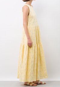 Une femme dans une robe florale jaune se tient de profil, mettant en valeur le design à volants et les motifs décoratifs de la robe, accompagnée de sandales légères.