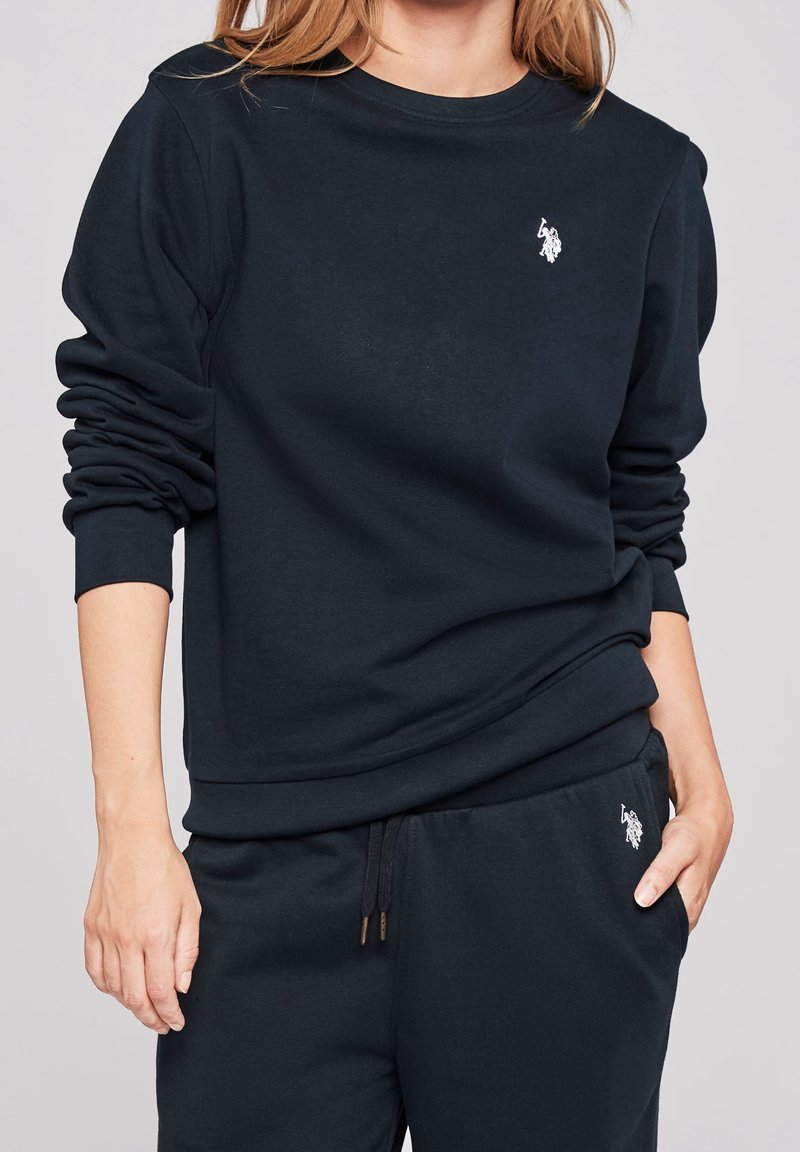 Femme portant un sweat-shirt noir à manches longues et un pantalon assorti avec un logo de joueur de polo blanc sur la poitrine et la cuisse, debout avec une main dans la poche.