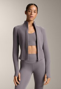 Ľahká, priliehavá atletická bunda v jemnej sivej farbe s vysokým golierom, plným zipsom a dlhými rukávmi, skombinovaná s bielo-zeleným športovým topom a legínami.