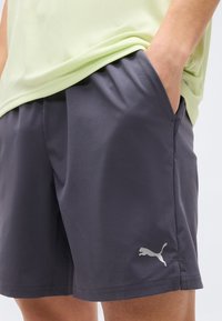 Άτομο που φοράει σκούρα γκρι αθλητικά σορτς Puma με τα χέρια στις τσέπες και ένα ανοιχτό πράσινο πουκάμισο ορατό από τη μέση και κάτω.