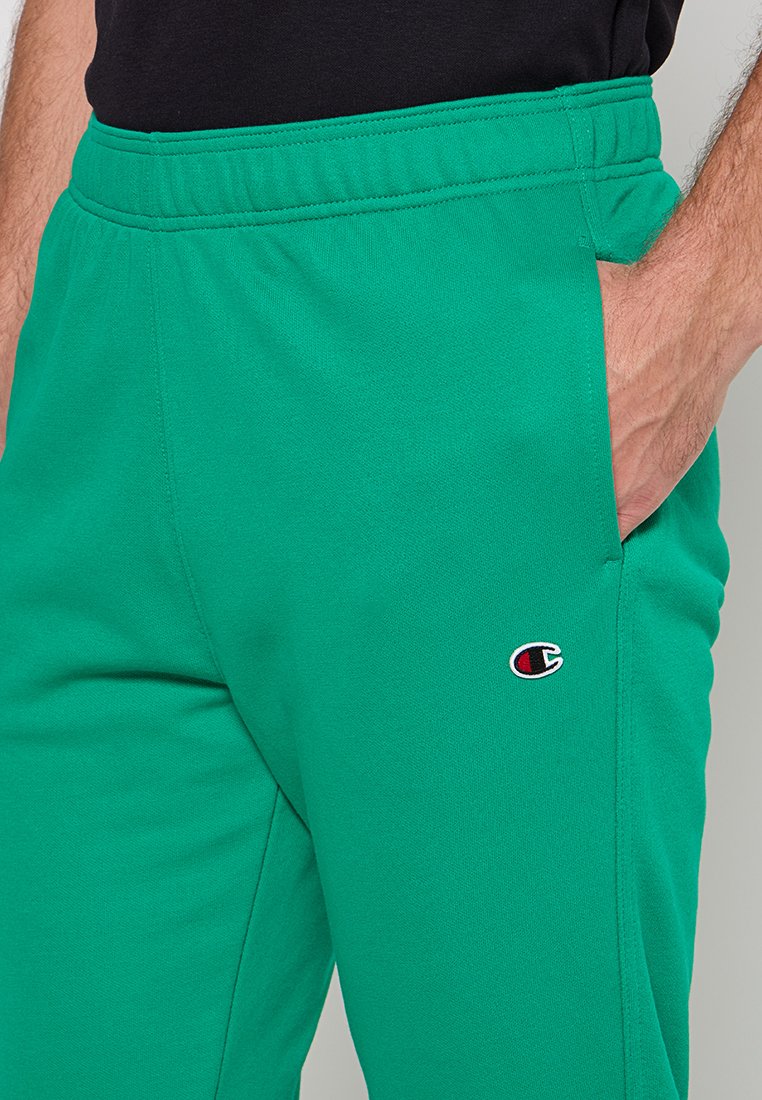 Pantalones de chándal verdes con cintura elástica, bolsillos laterales y pequeño parche de logo rojo y blanco en la parte superior de la pierna. Textura suave y diseño casual.