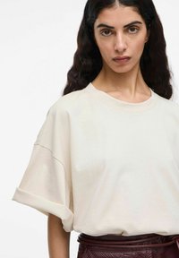 Kremo spalvos per didelė t-shirt su sukniukais, pagaminta iš minkštos medvilnės. Modelis dėvi blizgantį bordo spalvos gyvatės odos rašto sijoną.