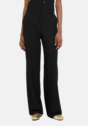 Donna che indossa pantaloni neri a gamba larga e scarpe piatte dorate, in piedi davanti a uno sfondo neutro.