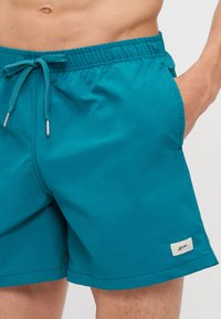 Shorts de bain teal en tissu léger, avec taille élastique et cordon de serrage réglable, ainsi que poches latérales. Petite étiquette de marque sur l'ourlet.