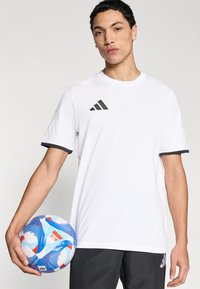 Λευκό κοντομάνικο t-shirt με μαύρες λεπτομέρειες, με ανάγλυφο μαύρο λογότυπο adidas. Το μοντέλο κρατά μια πολυχρωμη ποδοσφαιρική μπάλα.