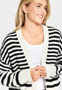 Cardigan en tricot rayé noir et blanc avec un col et des poignets côtelés, ouvert à l'avant et à coupe décontractée, porté par-dessus un top blanc.