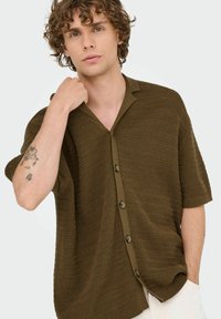 Brown knit short-sleeve shirt with a collar, featuring a textured pattern and five buttons down the front. Model poses casually.