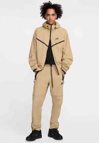 Beżowa kurtka z kapturem Nike z czarnymi akcentami, do tego pasujące beżowe spodnie z zamkami i sznurkami, noszone z czarnymi sneakersami.