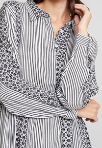 Blouse noire et blanche avec des rayures verticales et des accents à motifs. Col boutonné, manches longues, et coupe ample. Tissu texturé.