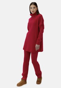 Röd ribbad stickad klänning med hög krage, kombinerad med matchande figurnära byxor och beige skor. Texturerat tyg och avslappnad passform.