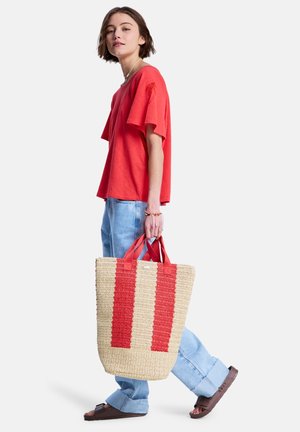 Mujer de cabello corto con camisa roja y jeans azul claro, llevando un bolso tote grande beige con rayas rojas, caminando con sandalias marrones.