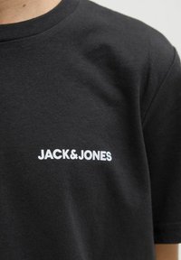 Crna pamučna majica s okruglim izrezom i kratkim rukavima. Na prednjoj strani nalazi se bijeli logotip s tekstom "JACK&JONES" centriran na prsima.
