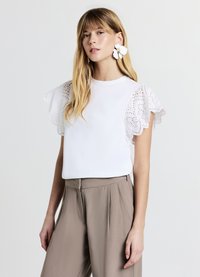 Haut blanc à manches courtes avec des accents en coton brodé et à volants. Associé à un pantalon beige taille haute. Design simple et élégant.
