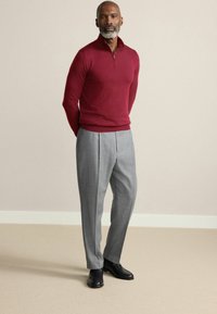 Pulover burgundy cu gât cu fermoar, pantaloni gri plisati și pantofi negri. Puloverul are un design pe corp; pantalonii sunt tăiați cu o textură moale.
