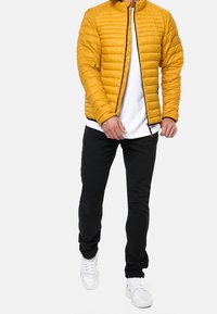 Gelbe, gestepfte Pufferjacke mit Stehkragen, Frontreißverschluss und schwarzen Akzenten, getragen über einem weißen Hemd und schwarzen Hosen. Weiße Sneakers.