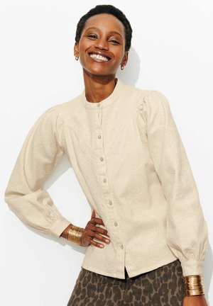 Femme souriante aux cheveux courts portant un chemisier crème texturé à boutons avec manches bouffantes et un pantalon imprimé léopard, main sur la hanche.