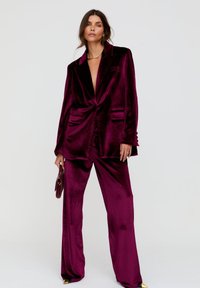 Samtanzug in tiefem Burgunderrot, mit locker sitzendem Blazer mit zwei Fronttaschen und weitem Hosenbein. Inklusive einer eleganten, kleinen Handtasche.